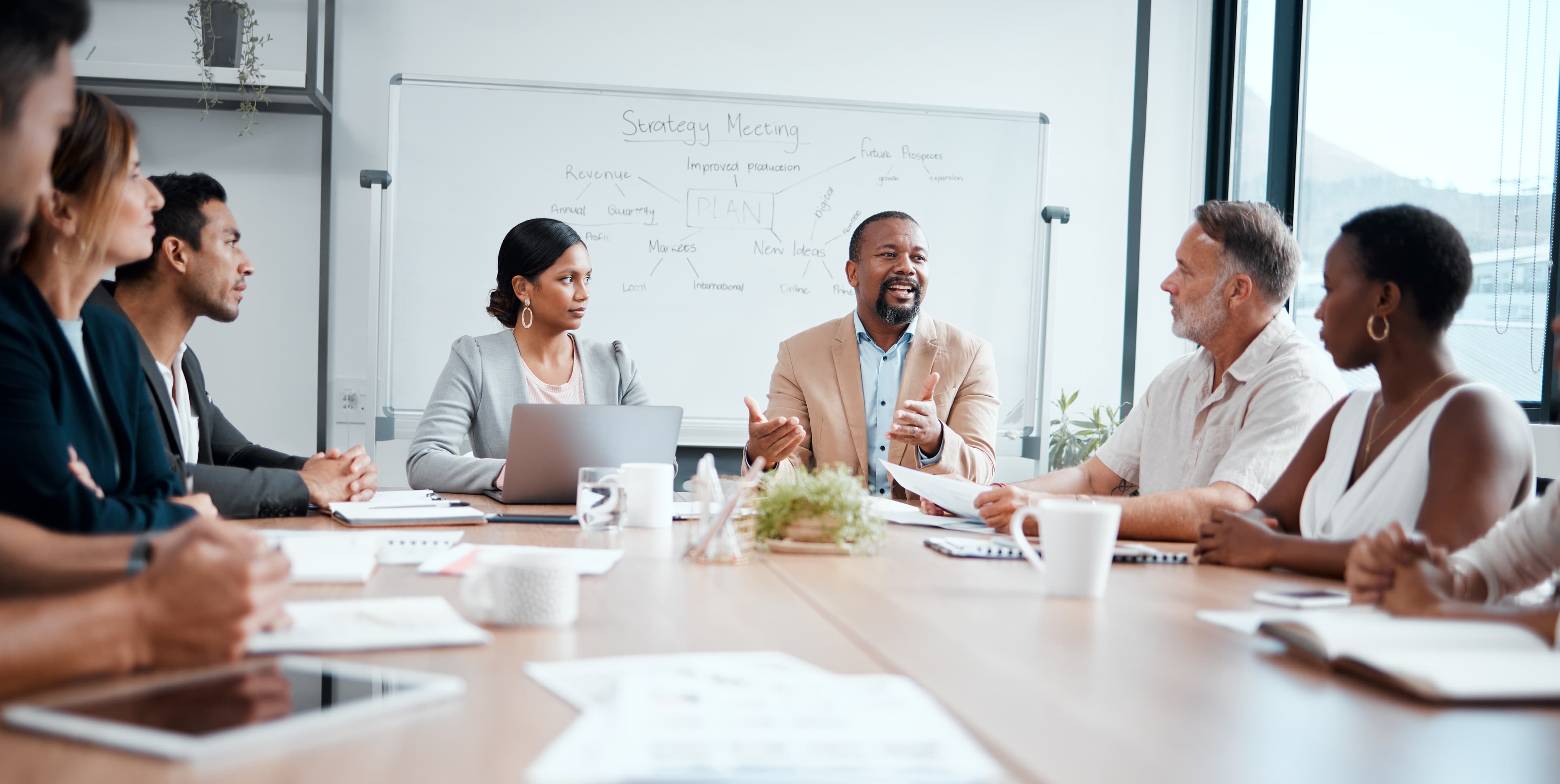 Diverse executive roundtable with seven or eight senior-level professionals sat at a meeting table having an interactive workshop