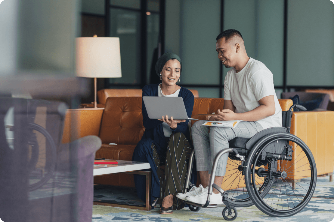 Two people, a woman seated on a couch next to a man using a wheelchair, are talking and looking at a computer